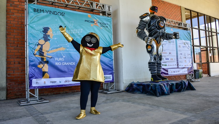 Imagem mostra mascote do evento na frente de banner oficial