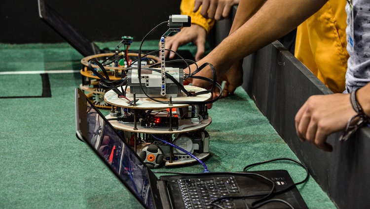 Imagem mostra, parcialmente, braços de competidores e, em foco, um robô sobre a plataforma de competição