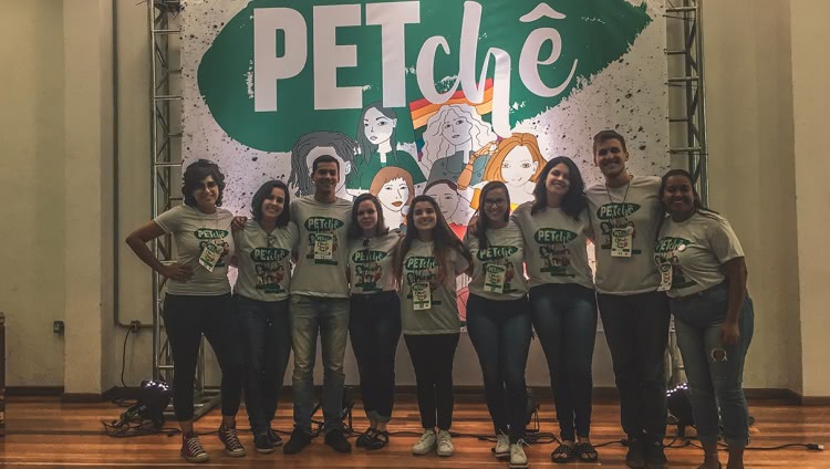 Na foto, nove alunos estão lado a lado com camisetas iguais, temáticas do evento. Eles estão abraçados em frente a um cartaz que tem as palavras “PETchê”. No desenho abaixo das palavras estão representadas várias pessoas. Atrás de uma das pessoas desenhadas está uma bandeira LGBT, com listras nas cores do arco-íris.