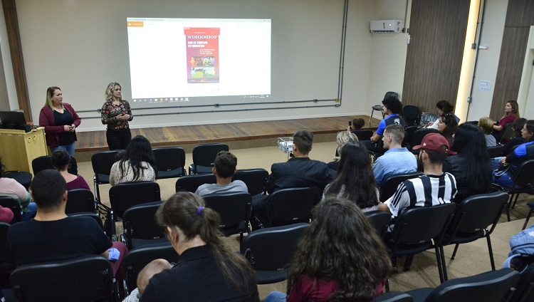 A foto mostra as organizadoras em frente a um projetor falando para uma plateia sentada em cadeiras em um auditório.