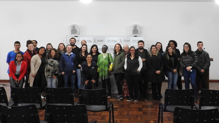 Na foto, aparece um grande número de pessoas, em pé, que posa para a foto. Eles estão numa sala com cadeiras. Atrás das pessoas em pé, há um banner com inscrições de textos da Prefeitura Municipal de Rio Grande