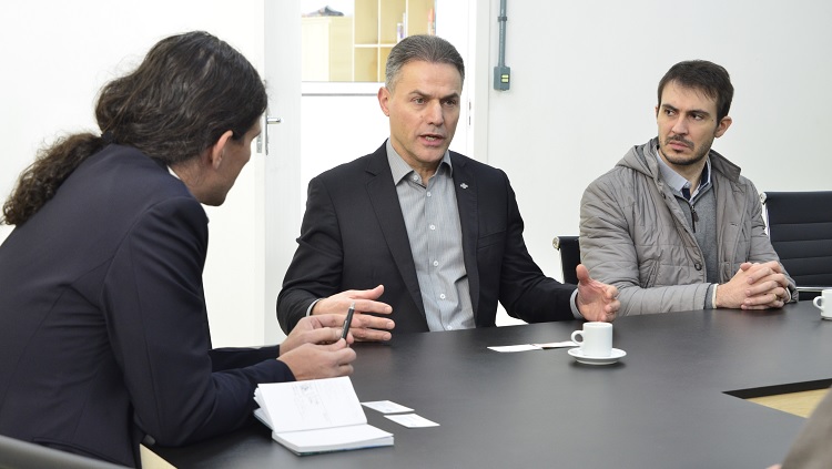 A foto mostra três homens em uma mesa de reuniões. O que está ao centro gesticula, enquanto os dois demais olham para ele.