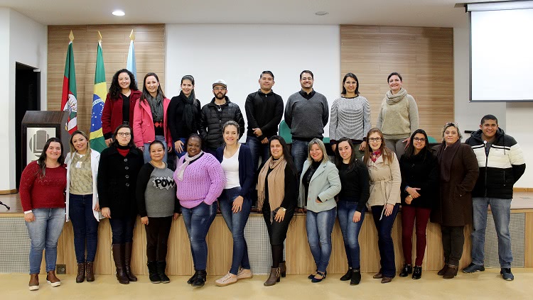 Na imagem aparece um grande grupo de pessoas, cerca de 20. Elas posam para a foto. Estão em um espaço de auditório. Metade do grupo está em cima de um palco, a outra metade está embaixo. Todos estão em pé. 