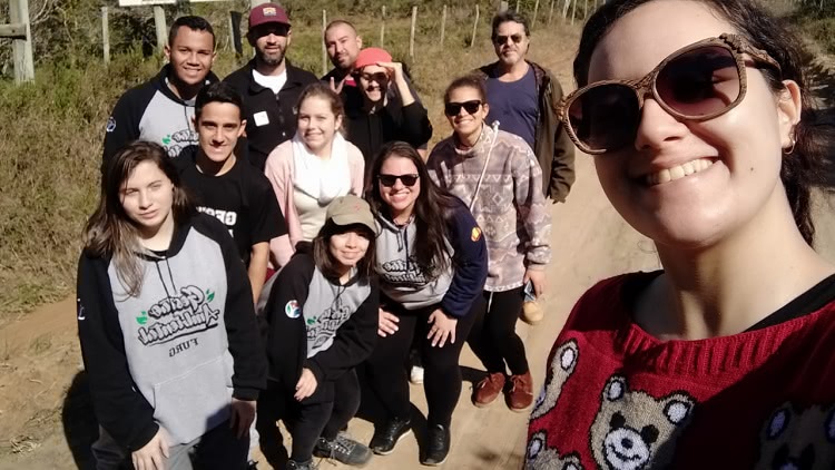 Uma jovem mulher, de óculos escuros aparece em primeiro plano. A foto é um selfie. Ao fundo, aparecem mais 11 pessoas. Todas posam para a foto. Estão em um local ao ar livre. Aparece estrada de terra e vegetação 