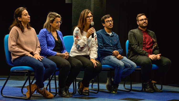 Mauren, idealizadora do projeto, aparece na foto em meio ao seu discurso, sentada segurando o microfone enquanto os demais componentes da mesa estão sentados ao seu lado.