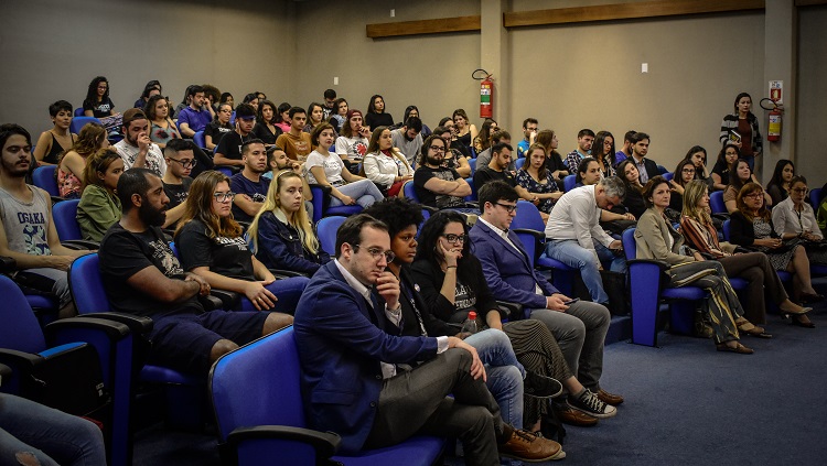 Imagem mostra várias pessoas sentadas nas cadeiras do auditório