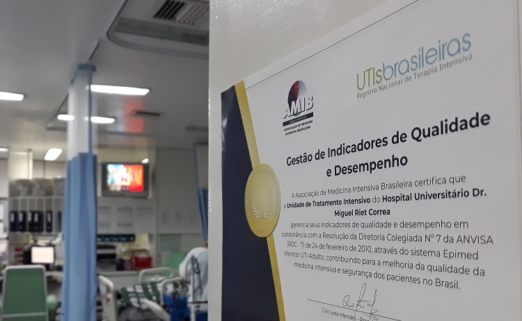 Um certificado aparece afixado a uma porta aberta, à direita da imagem O certificado é assinado por AMIB e UTIs Brasileiras. O título é Gestão de Indicadores de Qualidade e desempenho.   Pode-se ler, no certificado, que a Associação de Medicina Intensiva Brasileira certifica que a Unidade de Tratamento do Hospital Universitário Dr. Miguel Riet Correa gerencia seus indicadores de qualidade e desempenho em consonância com a Resolução da Diretoria Colegiada número 7 da ANVISA.  Ao fundo da porta que está aberta, é possível ver camas da UTI Adulto. 