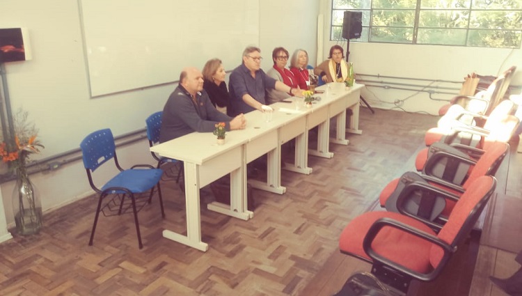 Na foto, os palestrantes da reunião estão sentados diante de uma mesa discursando para o público