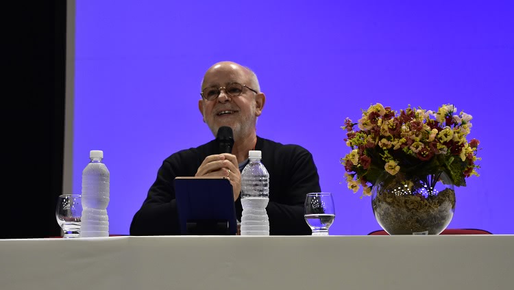 Imagem destaca homem, sentado à mesa, com microfone em punho