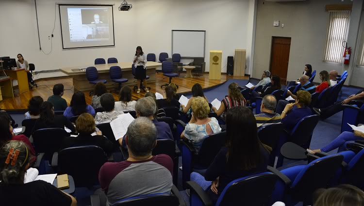 Em uma foto que mostra um auditório, o público aparece de costas. Ao fundo da imagem, no palco, a reitora Cleuza Maria Sobral Dias fala ao microfone.