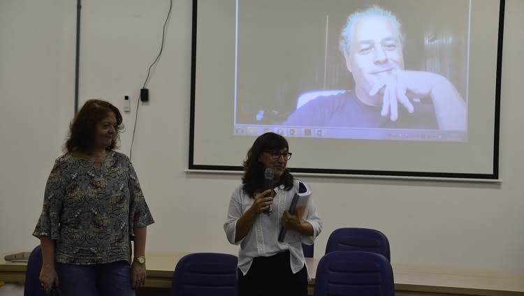 A presidente da CPA, Adriana Kivanski, e a reitora da FURG, Cleuza Maria Sobral Dias aparecem na foto. Elas estão no palco. Cleuza está com o microfone em uma mão e o relatório de avaliação institucional em outra. Atrás delas, em um telão, aparece um homem, que participa da reunião por teleconferência. 