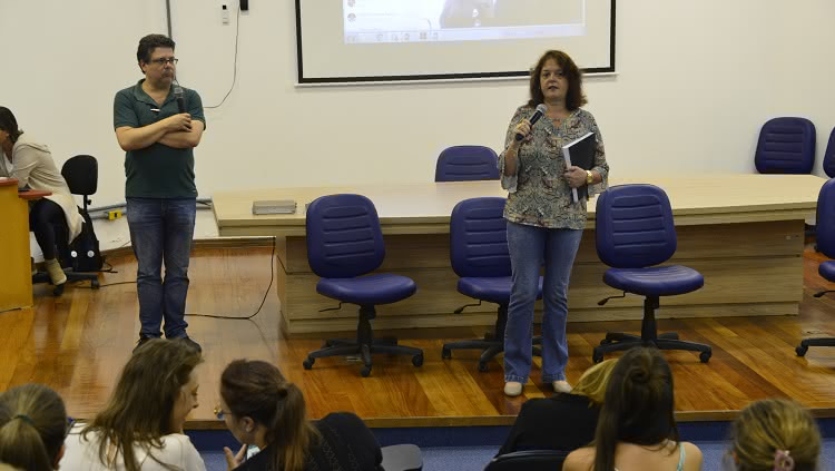 Um homem e uma mulher aparecem na foto. Eles estão no palco, ambos com microfone à mão. Ela parece estar falando e segura o relatório de avaliação institucional em uma das mãos. 