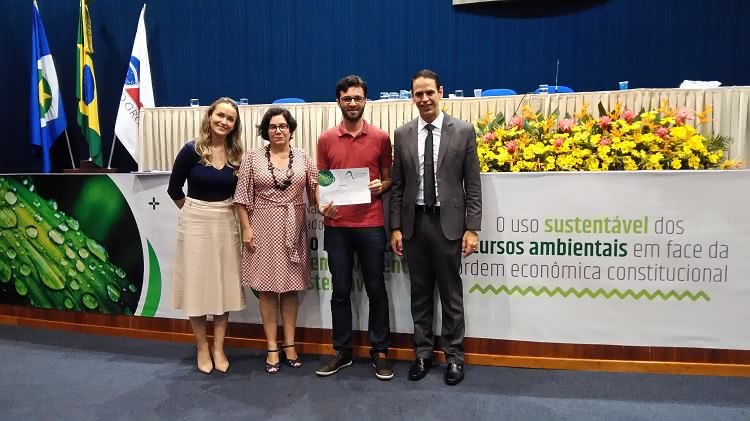 Na imagem, quatro pessoas aparecem em frente a uma mesa em um espaço que parece ser um auditório. Elas estão uma ao lado da outra e olham para a foto. À direita duas mulheres, ao lado esquerdo delas o mestrando premiado e, ao lado dele, um homem. Atrás deles, na faixa que está na mesa do evento, é possível ler parte de um texto, que diz: “o uso sustentável dos recursos ambientais em face da ordem econômica constitucional”. 