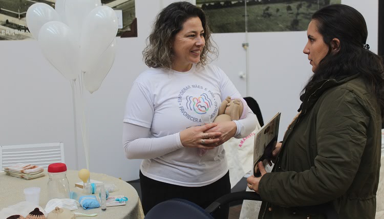 Na foto, duas mulheres aparecem conversando. Uma delas está com uma camiseta em que consta o texto: "empoderar mães e pais. Favorecer a amamentação". Esta mulher segura nos braços um bebê de pano e sorri. A outra, de frente para ela, a observa. Ao lado delas há uma mesa com materiais de apoio para práticas de amamentação. Na mesa, também há balões de cor branca. 