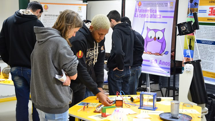 Cerca de seis jovens rapazes aparecem na foto. Quatro estão de costas, dois estão de frente, diante de uma mesa, em que testam equipamentos. Ao fundo, atrás deles, aparece um banner em que se lê: "Licenciatura em Matemática"