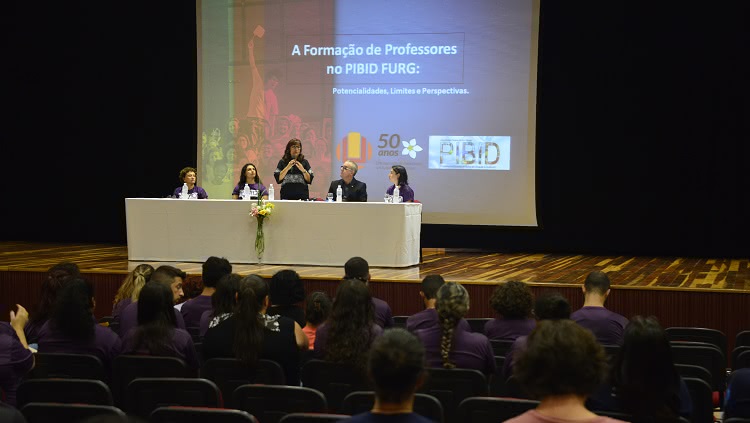 A foto mostra um auditório. No palco, que está iluminado, quatro pessoas estão sentadas atrás de uma mesa. Uma quinta pessoa, ao centro, está em pé e com o microfone à mão. No telão, ao fundo, o texto: 