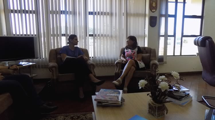 A foto mostra a prefeita de São José do Norte, Fabiany Zogbi Roig, e a reitora da FURG, Cleuza Maria Sobral Dias. Elas estão sentadas em poltronas, no gabinete da reitora e a situação é de conversa. À frente delas, uma mesa em que aparecem revistas e livros e um arranjo de flores e, ao fundo, janelas de vidro com persianas entre abertas. 