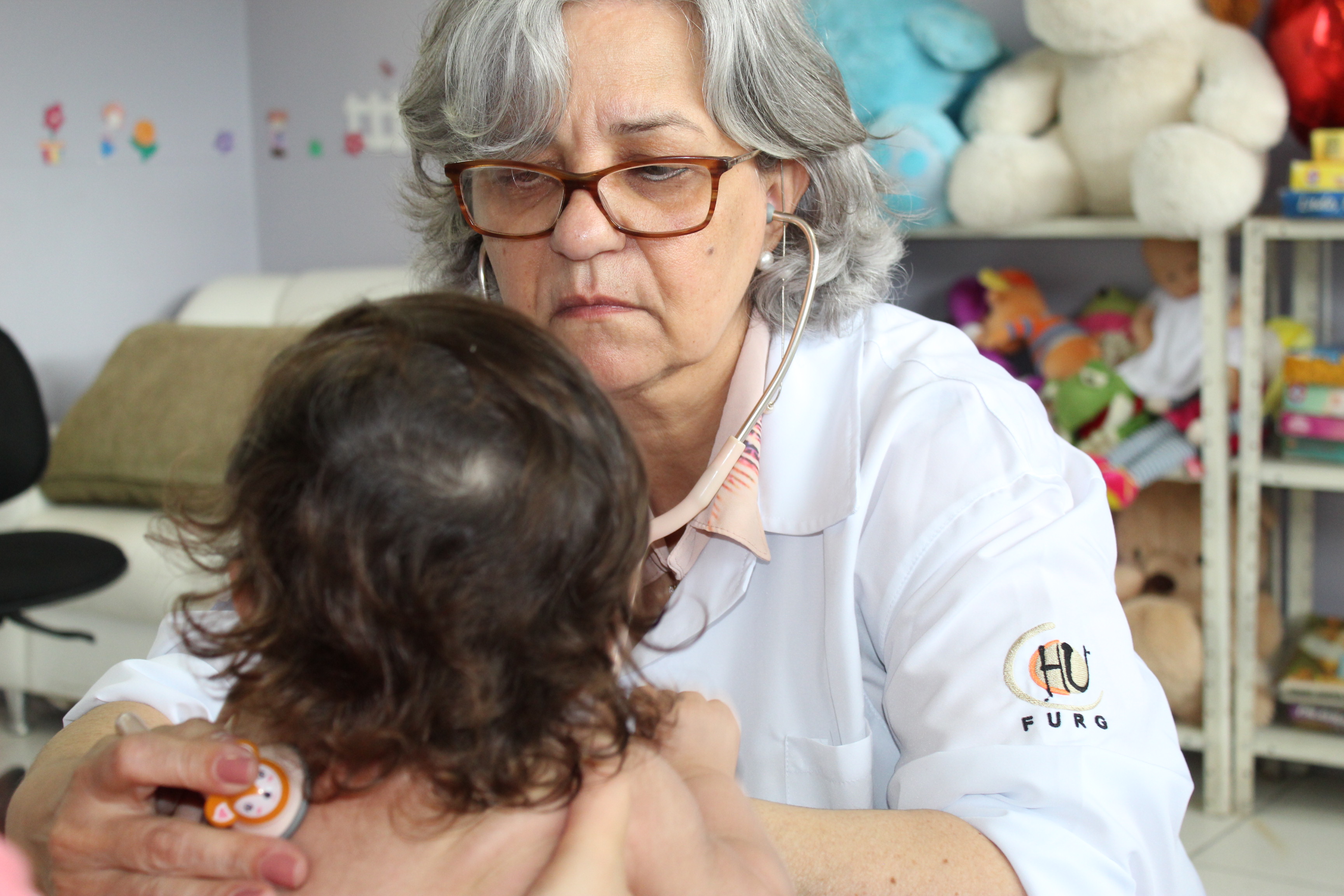 A foto mostra uma médica atendendo a uma criança. A médica aparece de frente para a câmera, e a criança de costas. Ao fundo os elementos são de um quarto hospitalar infantil. Há ursos de pelúcia em uma estante e adesivos coloridos na parede. A médica usa um estetoscópio, que está nas costas da criança. Sua feição é de concentração. 