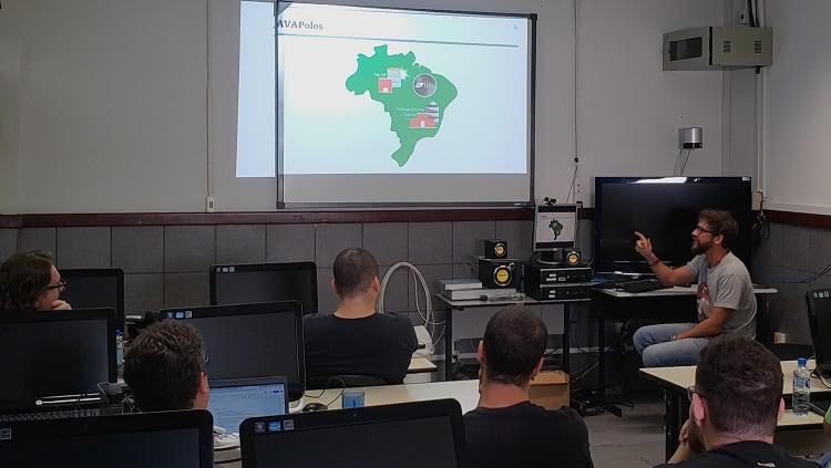 A foto mostra um laboratório de informática. Há algumas pessoas que aparecem de costas, diante de computadores desligados. Ao fundo, um mapa do Brasil é projetado numa lousa branca. Um homem, que está sentado numa carteira de professor, aponta para o mapa projetado. No mapa há dados ilegíveis e, acima, consta a descrição: AVAPolos. 