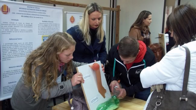 Três estudantes, duas mulheres e um homem, fazem demonstração de um experimento científico com garrafas PET em um evento acadêmico. Eles estão no entorno de uma mesa. À frente, uma mulher observa. Atrás deles, há dois pôsteres de apresentação de trabalhos científicos, ambos com a marca da FURG. 