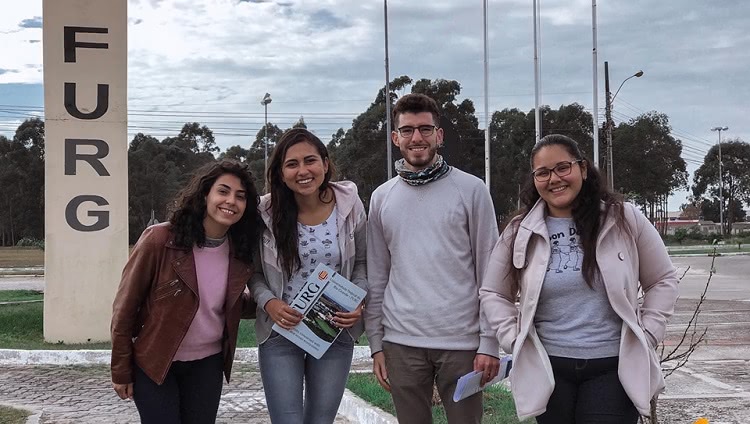 A foto mostra quatro jovens estudantes em área externa da FURG. São quatro jovens mulheres, um jovem homem. Uma das mulheres segura uma pasta com o nome da FURG. Ao fundo, atrás deles, há a bandeira da universidade, em mastro, a do Brasil e a do Rio Grande do Sul, e um pilar em que está escrito FURG. 