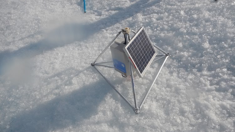 Na imagem, um tripé de metal sobre uma superfície de gelo, com uma placa de luz solar