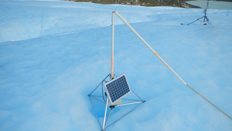 Sobre uma superfície de gelo, aparece um tripé de metal com um painel de luz solar. No centro dele, há segmentos de metal, que se desdobram no chão, como hastes de barracas