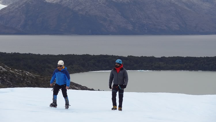 Dois homens agasalhados, com calçados com garras para neve, caminham sobre o gelo. Eles também usam capacete. Ao fundo deles se vêem um lago e, mais atrás, montanhas