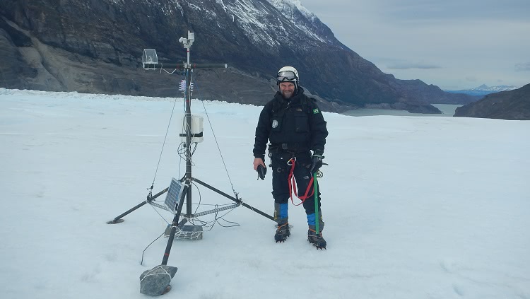 Um homem aparece de corpo inteiro, bem agasalhado, com grandes óculos, capacete e botas com garras para neve aparece sobre uma geleira, ao lado de um equipamento. O equipamento é um pouco maior que ele, em altura, e se sustenta sobre um tripé. 