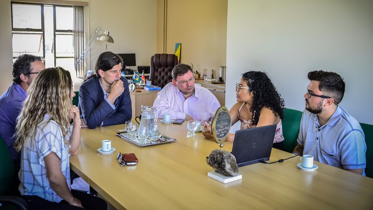 Imagem mostra seis pessoas, duas mulheres e quatro homens ao redor de uma mesa de reuniões. No centro, um notebook, usado para uma apresentação de slides. Os semblantes revelam interesse por parte dos rostos que estão virados para uma das mulheres, que, em contrapartida, assume postura de quem está comunicando sobre algo