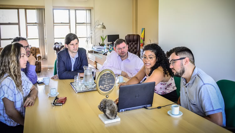 Imagem mostra seis pessoas, duas mulheres e quatro homens ao redor de uma mesa de reuniões. No centro, um notebook, usado para uma apresentação de slides. Os semblantes revelam interesse por parte dos rostos que estão virados para uma das mulheres, que, em contrapartida, aponta para o notebook 