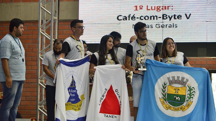 Quatro pessoas estão num palco, dois rapazes e duas meninas. Eles seguram bandeiras, uma do colégio Afonso Pena, outro do Estado de Minas Gerais, e outra do município de São Tiago. Ao fundo, num telão, a indicação de primeiro lugar. 