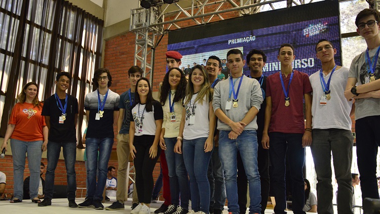 Na foto aparecem 14 pessoas, posicionadas lado a lado, em frente a um painel de led. Todas estão com medalhas nos pescoços.