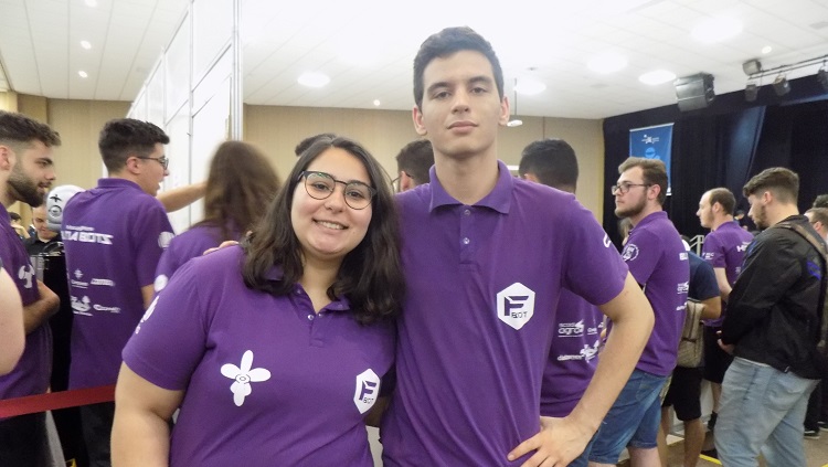 Na foto, uma jovem mulher e um rapaz posam. Eles estão com camisetas iguais, de cor roxa. Atrás deles há pessoas vestindo a mesma camiseta. 