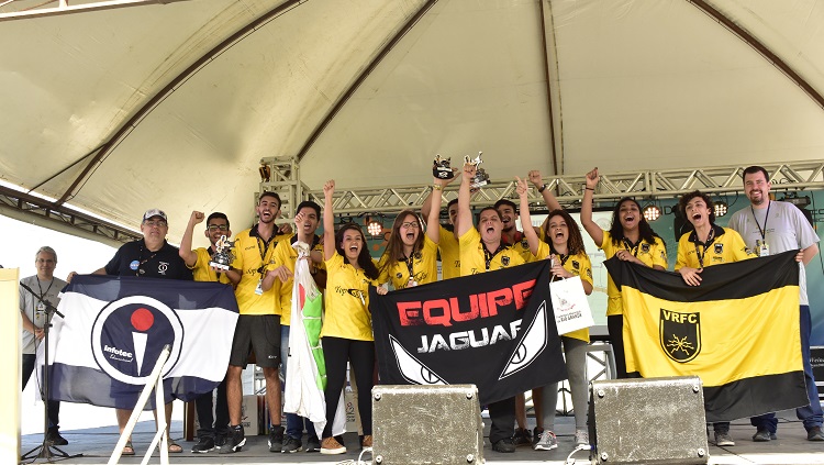 Uma equipe com mais de 20 pessoas está num palco. Seguram um cartaz com o nome ITA