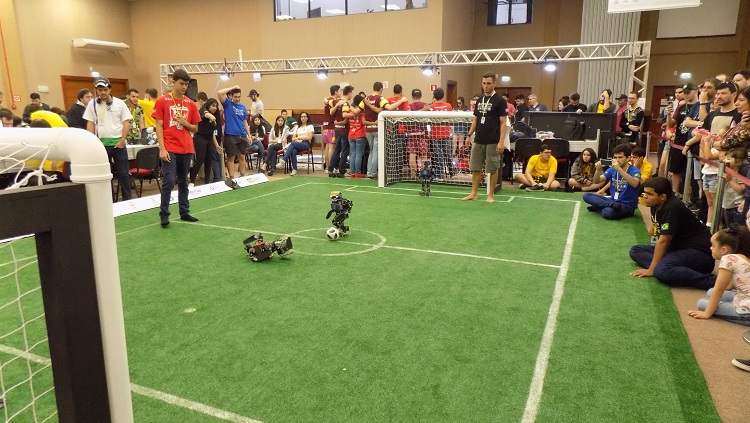 Na imagem há um campo de futebol e robôs jogando