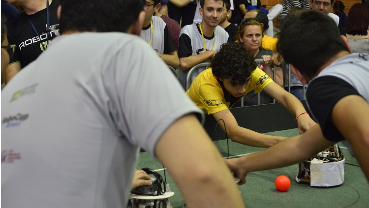 Imagem foca competidor ajustando um robô na pista de competição, ele traja uma camiseta na amarela. Em primeiro plano, dois jovens de costas aparecem desfocado