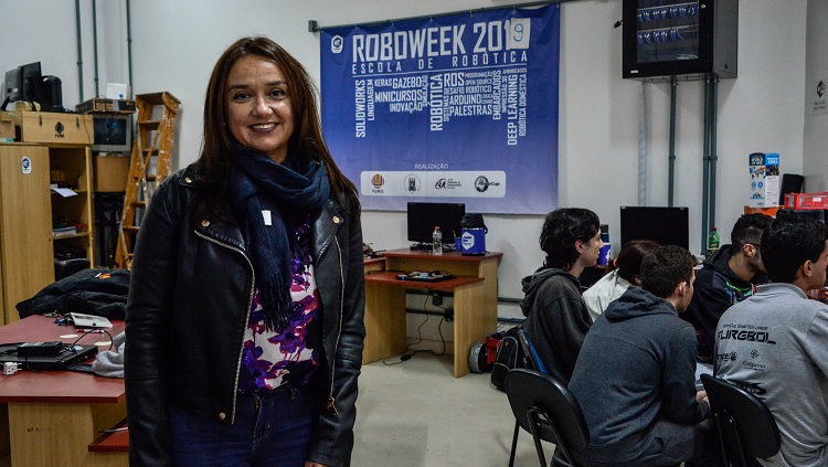 Em primeiro plano, aparece uma mulher em pé em uma ampla sala de aula. Ao fundo, na parede, há um banner, em que está escrito Roboweek 2019 - escola de robótica. Há mesas e cadeiras ao fundo. Cerca de 5 estudantes aparecem sentados em uma mesa, de costas para a imagem.