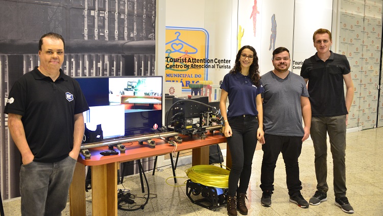 No centro da foto há uma mesa retangular e sobre ela estão equipamentos eletrônicos. Ao lado direito da mesa, uma moça e dois rapazes posam para a foto. Ao lado esquerdo, outro rapaz. 