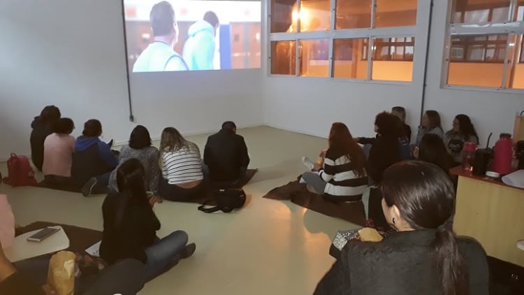 Em uma sala, cerca de 20 estudantes aparecem sentados sobre mantas no chão, em pequenos grupos. Ao fundo, há um filme sendo projetado em uma parede branca.