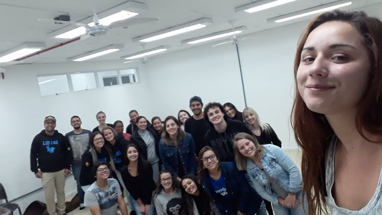 A foto é uma selfie. Uma menina sorridente aparece em primeiro plano. Ao fundo, 21 pessoas, na maior parte jovens, posam para a foto. 