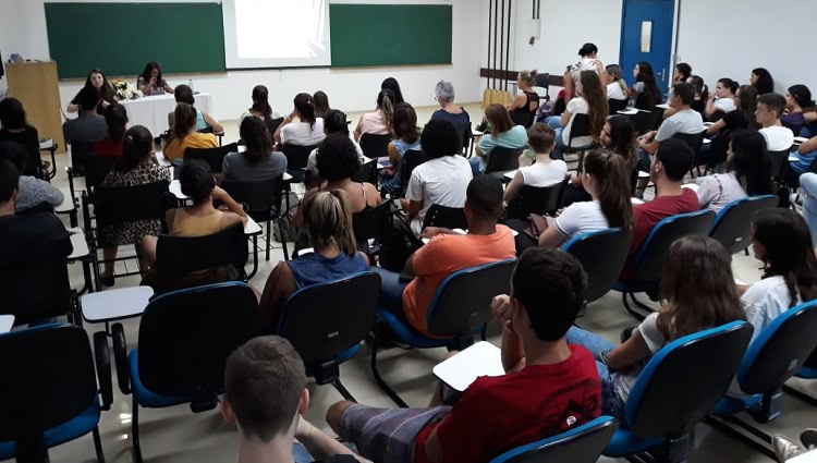 a foto mostra um auditório cheio. Ao fundo da imagem, em uma mesa, aparecem duas mulheres sentadas atrás de uma mesa. Ao fundo delas, ao lado direito, uma tela de projetor. Uma das mulheres na mesa fala ao microfone. 