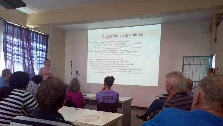 Em uma sala de aula, aparecem cerca de 10 pessoas. Elas estão sentadas às classes, de costas na foto, e olham para uma mulher que está em pé, à frente do quadro e gesticula. Atrás dela, numa tela, há uma projeção com planilhas e o texto: “Sugestão de planilhas”. 