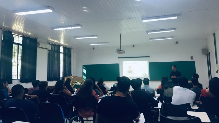 Sala de aula com pessoas olhando para frente em primeiro plano. Na frente homem em pé, atrás dele quadro verde, na lateral duas janelas com cortinas.