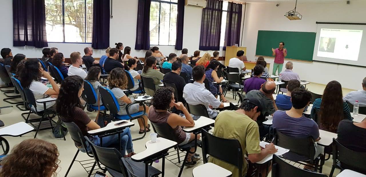 A foto mostra uma sala de aula cheia. Na frente da turma, um professor fala aos alunos com uma projeção ao fundo.