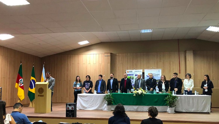 Grupo de pessoas reunidas lado a lado atrás de uma mesa. Uma pessoa no púlpito com microfone na mão.