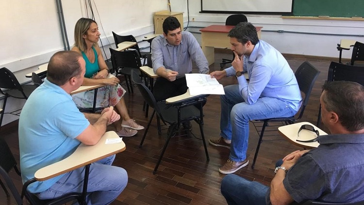 Em uma sala de aula, estão sentados em círculo, quatro homens e uma mulher. Dois deles se detém sobre um documento.