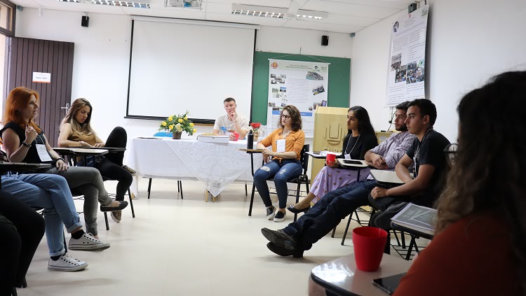 a imagem mostra uma sala de aula. Os alunos estão dispostos em círculo, sentados em cadeiras. Ao fundo, aparece uma mesa coberta com uma toalha branca e sobreposta por uma toalha de renda. Em cima da mesa há um arranjo de flores. Um rapaz está sentado atrás desta mesa. 