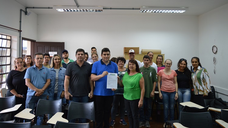 Em uma sala de aula, cerca de 20 pessoas estão reunidas e pousam para a foto. À frente do grupo estão o pró-reitor Eduardo Saldanha Vogelmann e a vereadora Marcia Lucas. Eles seguram juntos um documento. 