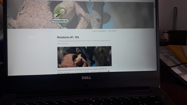 A foto é de um computador estilo notebook. O cenário está escuro e é possível apenas ver que a tela mostra um site em que no topo aparece o texto Relatório #1: RS e, abaixo, a imagem de um pássaro João de Barro em frente a seu ninho